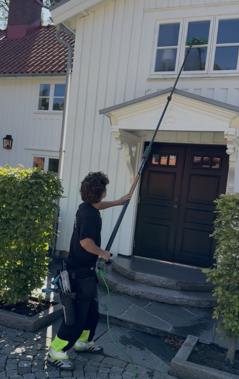 Fönsterputsare från SeBra Group tvättar ett fönster på övervåningen med vattenmatad teleskopstång, framför ett vitt trähus i sommardagsljus. Verktygsbälte och slang syns.